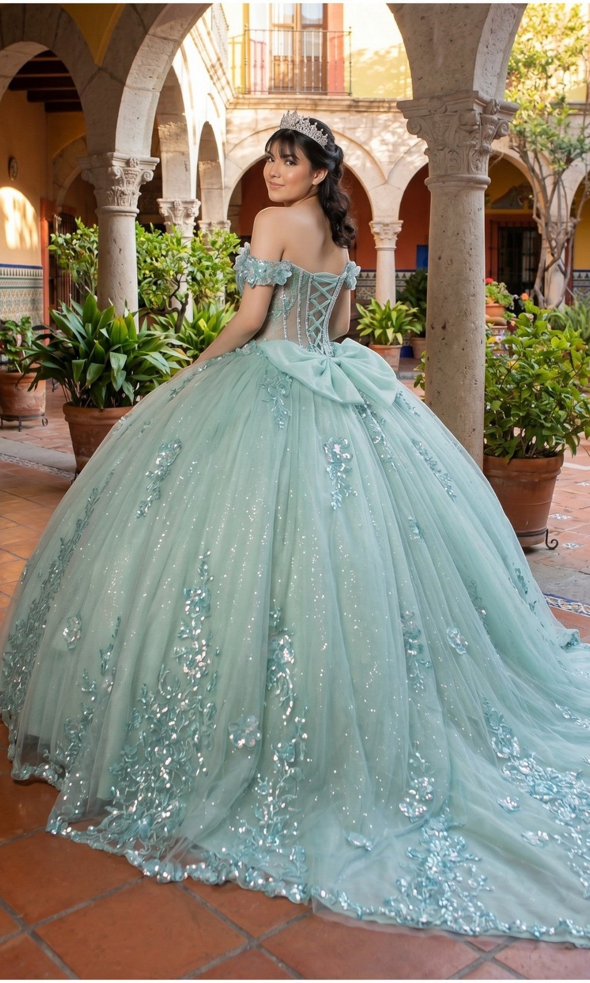 A young woman stands in a courtyard, looking back over her shoulder, wearing the Quinceanera Dress 8370J By Cinderella Couture—a light blue ball gown featuring floral appliques and an illusion corset bodice.